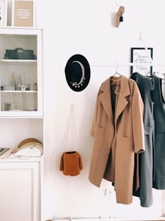 A neatly arranged hallway with a coat rack features a beige overcoat and a grey dress hanging. Next to them, a black hat with decorative elements is placed. A small rust-colored tassel bag hangs beneath the coat. Above a cabinet, a minimalistic shelf displays some decorative items and framed art, alongside various small objects. The overall setting is minimalistic and organized, with a neutral color palette.