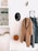 A neatly arranged hallway with a coat rack features a beige overcoat and a grey dress hanging. Next to them, a black hat with decorative elements is placed. A small rust-colored tassel bag hangs beneath the coat. Above a cabinet, a minimalistic shelf displays some decorative items and framed art, alongside various small objects. The overall setting is minimalistic and organized, with a neutral color palette.
