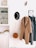 A neatly arranged hallway with a coat rack features a beige overcoat and a grey dress hanging. Next to them, a black hat with decorative elements is placed. A small rust-colored tassel bag hangs beneath the coat. Above a cabinet, a minimalistic shelf displays some decorative items and framed art, alongside various small objects. The overall setting is minimalistic and organized, with a neutral color palette.