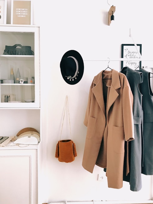 A neatly arranged hallway with a coat rack features a beige overcoat and a grey dress hanging. Next to them, a black hat with decorative elements is placed. A small rust-colored tassel bag hangs beneath the coat. Above a cabinet, a minimalistic shelf displays some decorative items and framed art, alongside various small objects. The overall setting is minimalistic and organized, with a neutral color palette.