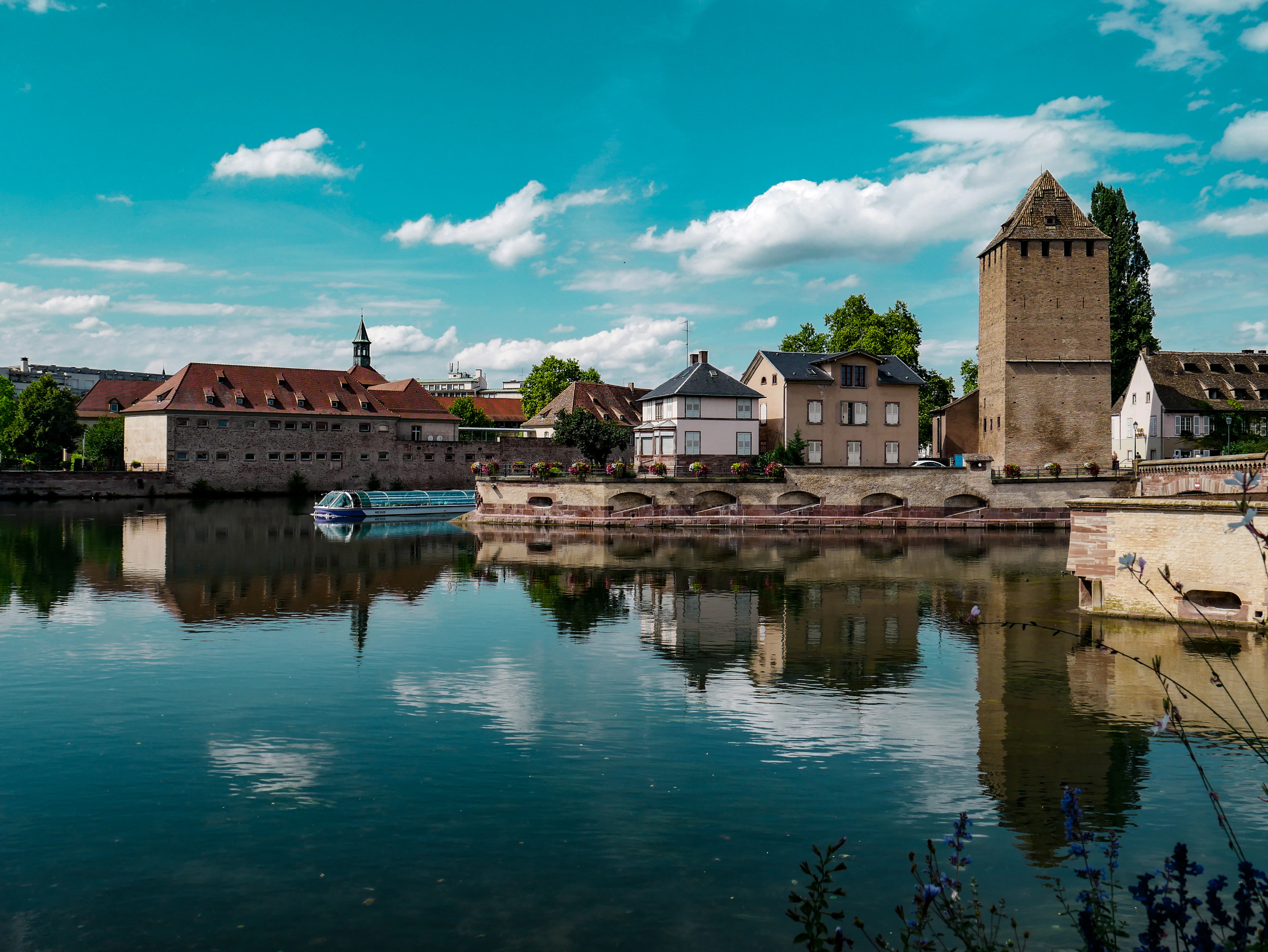 A medieval stone bridge tower and the long arcaded weir of the Barrage Vauban mirrored in the still River Ill at Strasbourg, old-town roofs behind under a summer sky of white cloud.