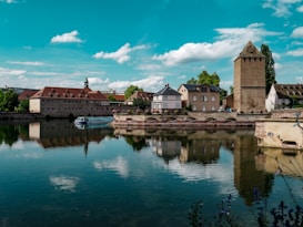 A serene riverside scene featuring several historic buildings with red-tiled roofs, reflected in the calm waters below. A tower stands prominently on the right, while a tour boat is visible on the water. The sky is partly cloudy with scattered blue above, and lush greenery surrounds the area.