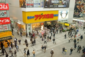 A busy shopping area with numerous people walking and standing near a brightly colored store. The storefront features large advertisements, posters of anime characters, and merchandise displays. The area is bustling with activity, and there are additional shops and signs visible in the surrounding area.