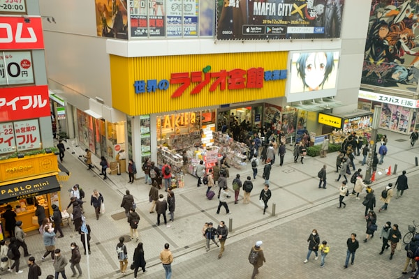 A busy shopping area with numerous people walking and standing near a brightly colored store. The storefront features large advertisements, posters of anime characters, and merchandise displays. The area is bustling with activity, and there are additional shops and signs visible in the surrounding area.