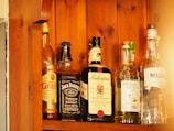 Several bottles of alcohol are lined up on a wooden shelf. Each bottle has a distinct label, indicating different types of spirits including whiskey and gin. The wood grain of the shelf adds a warm, rustic look to the image.