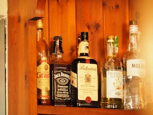Several bottles of alcohol are lined up on a wooden shelf. Each bottle has a distinct label, indicating different types of spirits including whiskey and gin. The wood grain of the shelf adds a warm, rustic look to the image.