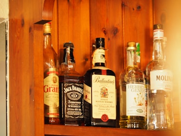 Several bottles of alcohol are lined up on a wooden shelf. Each bottle has a distinct label, indicating different types of spirits including whiskey and gin. The wood grain of the shelf adds a warm, rustic look to the image.