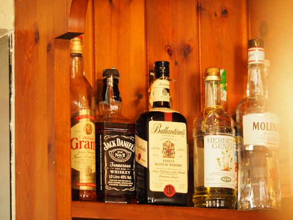 Several bottles of alcohol are lined up on a wooden shelf. Each bottle has a distinct label, indicating different types of spirits including whiskey and gin. The wood grain of the shelf adds a warm, rustic look to the image.