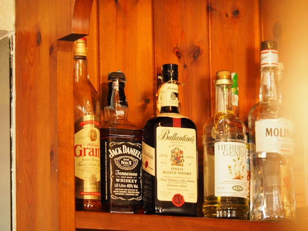 Several bottles of alcohol are lined up on a wooden shelf. Each bottle has a distinct label, indicating different types of spirits including whiskey and gin. The wood grain of the shelf adds a warm, rustic look to the image.