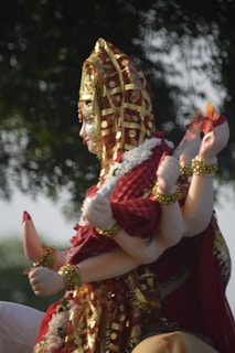 A statue of a multi-armed deity adorned with intricate gold and red garments. The figure holds various objects in its hands, and the background features blurred greenery.