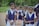 Several young students dressed in school uniforms stand outdoors on a dirt path, surrounded by greenery. The children wear navy blue vests and white shirts, each carrying a backpack.