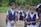 Several young students dressed in school uniforms stand outdoors on a dirt path, surrounded by greenery. The children wear navy blue vests and white shirts, each carrying a backpack.