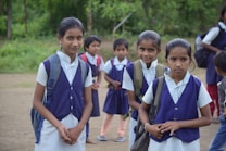 Several young students dressed in school uniforms stand outdoors on a dirt path, surrounded by greenery. The children wear navy blue vests and white shirts, each carrying a backpack.