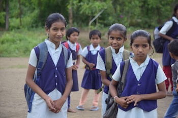 Several young students dressed in school uniforms stand outdoors on a dirt path, surrounded by greenery. The children wear navy blue vests and white shirts, each carrying a backpack.