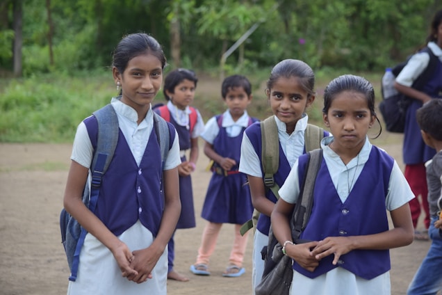 Several young students dressed in school uniforms stand outdoors on a dirt path, surrounded by greenery. The children wear navy blue vests and white shirts, each carrying a backpack.
