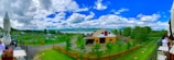 A scenic landscape featuring a barn with a red and yellow exterior surrounded by green trees and a wooden fence. In the background, there's a large lake or river with hills and a cloudy blue sky above. There are outdoor seating areas with umbrellas and tables to the sides.