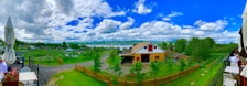 A scenic landscape featuring a barn with a red and yellow exterior surrounded by green trees and a wooden fence. In the background, there's a large lake or river with hills and a cloudy blue sky above. There are outdoor seating areas with umbrellas and tables to the sides.