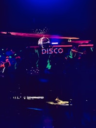 A dimly lit room featuring colorful neon lights spelling out 'disco'. There is a disco ball reflecting light across the ceiling, creating a lively and vibrant atmosphere. The room is filled with abstract patterns and a subdued ambiance.