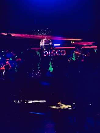 A dimly lit room featuring colorful neon lights spelling out 'disco'. There is a disco ball reflecting light across the ceiling, creating a lively and vibrant atmosphere. The room is filled with abstract patterns and a subdued ambiance.