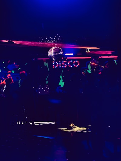 A dimly lit room featuring colorful neon lights spelling out 'disco'. There is a disco ball reflecting light across the ceiling, creating a lively and vibrant atmosphere. The room is filled with abstract patterns and a subdued ambiance.