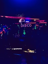 A dimly lit room featuring colorful neon lights spelling out 'disco'. There is a disco ball reflecting light across the ceiling, creating a lively and vibrant atmosphere. The room is filled with abstract patterns and a subdued ambiance.