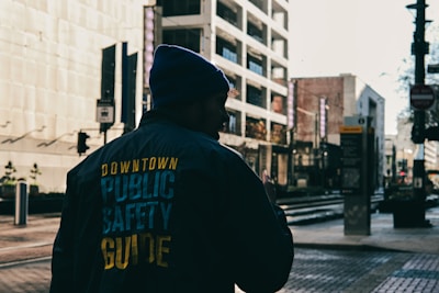 A person dressed in a jacket and beanie, with the words 'Downtown Public Safety Guide' on the back, walks down an urban street. The scene includes modern buildings and street elements like traffic lights and signs. Sunlight casts long shadows and highlights architectural details.