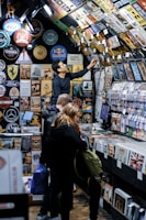 A bustling vintage shop displays a vast collection of retro signs, posters, and memorabilia on its walls and ceiling. Several customers browse through vinyl records and nostalgic items, with one person reaching up to examine a sign. The setting has a cozy, nostalgic feel, with items organized neatly on shelves.