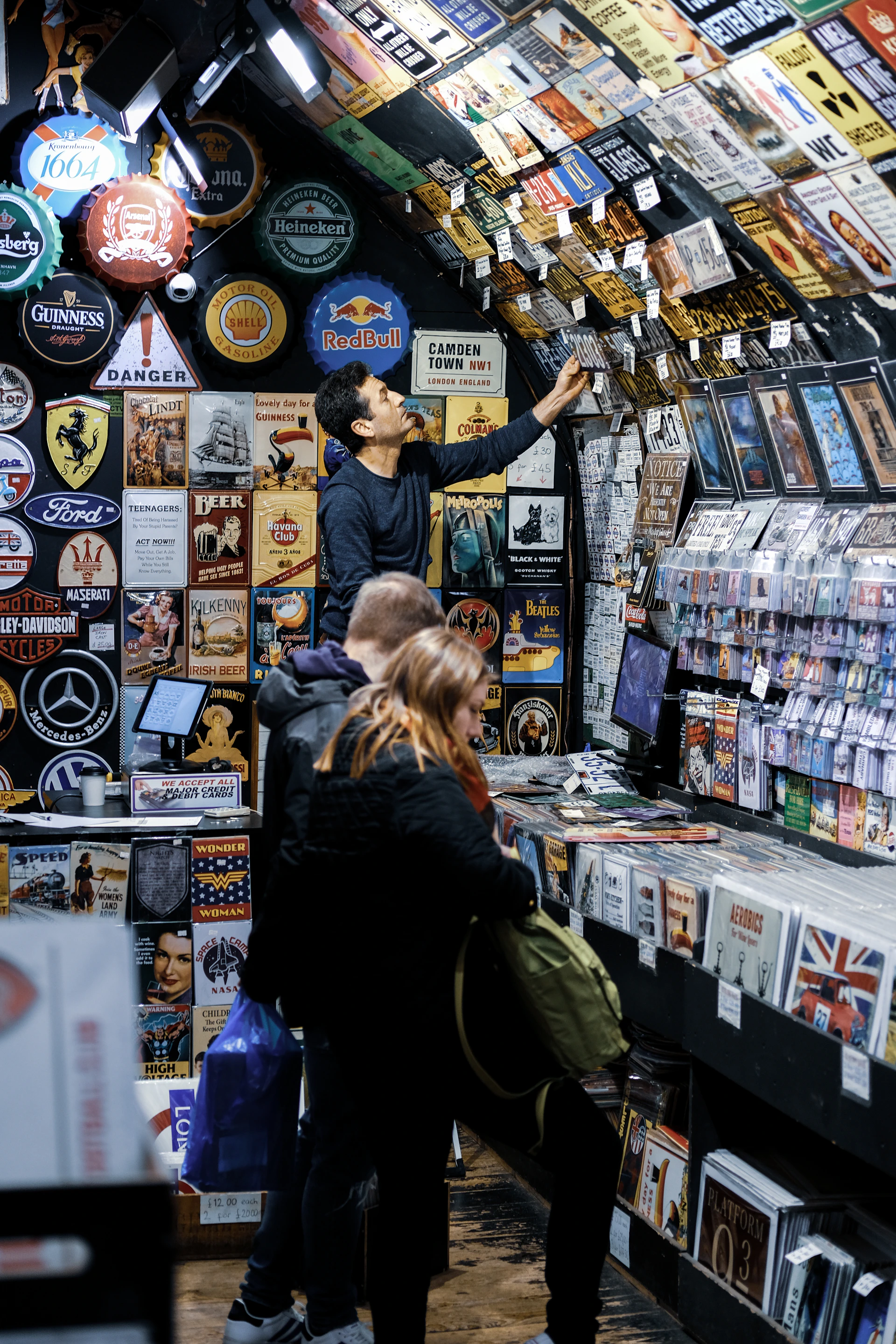 A bustling vintage shop displays a vast collection of retro signs, posters, and memorabilia on its walls and ceiling. Several customers browse through vinyl records and nostalgic items, with one person reaching up to examine a sign. The setting has a cozy, nostalgic feel, with items organized neatly on shelves.