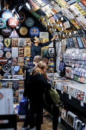 A bustling vintage shop displays a vast collection of retro signs, posters, and memorabilia on its walls and ceiling. Several customers browse through vinyl records and nostalgic items, with one person reaching up to examine a sign. The setting has a cozy, nostalgic feel, with items organized neatly on shelves.