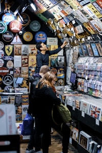 A bustling vintage shop displays a vast collection of retro signs, posters, and memorabilia on its walls and ceiling. Several customers browse through vinyl records and nostalgic items, with one person reaching up to examine a sign. The setting has a cozy, nostalgic feel, with items organized neatly on shelves.