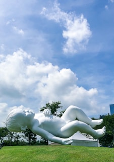 A large white sculpture of a baby is positioned on a grassy hill under a bright blue sky with scattered clouds. The sculpture depicts the baby in a reclining pose, surrounded by a natural setting with trees in the background and a modern cityscape peeking through.