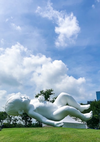 A large white sculpture of a baby is positioned on a grassy hill under a bright blue sky with scattered clouds. The sculpture depicts the baby in a reclining pose, surrounded by a natural setting with trees in the background and a modern cityscape peeking through.