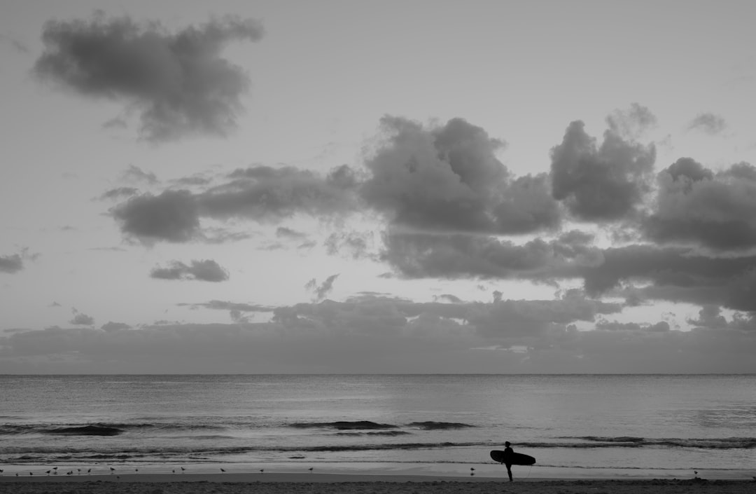 grayscale photography of person standing while holding surfboard viewing body of water