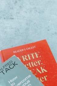 Two books are arranged on a surface, with one book titled 'WRITE Better, SPEAK Better' in red and the other titled 'How to Overcome Your Nervous Tension' by Alfred Tack in a muted green color.
