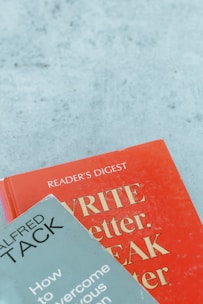 Two books are arranged on a surface, with one book titled 'WRITE Better, SPEAK Better' in red and the other titled 'How to Overcome Your Nervous Tension' by Alfred Tack in a muted green color.