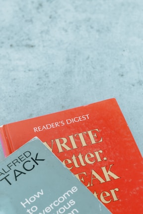 Two books are arranged on a surface, with one book titled 'WRITE Better, SPEAK Better' in red and the other titled 'How to Overcome Your Nervous Tension' by Alfred Tack in a muted green color.