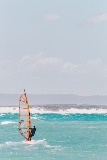 A person engages in windsurfing on a vibrant turquoise sea with whitecaps, under a clear blue sky. The windsurfer is balancing skillfully, holding onto the sail which is brightly colored in orange and yellow. In the background, distant hilly landforms are visible, with a hazy appearance.