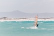 A windsurfer glides across turquoise ocean waters, with waves splashing around them. The background features misty, mountainous terrain and sandy shores.