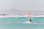A windsurfer glides across turquoise ocean waters, with waves splashing around them. The background features misty, mountainous terrain and sandy shores.
