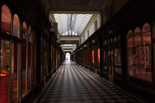 A long, narrow shopping arcade with checkered tile flooring and a glass ceiling that lets in natural light. The walls are lined with shop windows displaying various merchandise. The space has an elegant, vintage feel, enhanced by the warm lighting from the shopfronts.