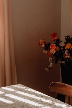 A cozy interior scene with a wooden table covered by a lace tablecloth. In the background, a bouquet of colorful flowers adds a touch of warmth. Soft natural light gently filters through beige curtains, casting shadows on the table.
