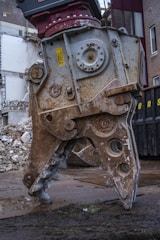 A large, rusted metal demolition claw is positioned on a wet, muddy ground, with crumbling bricks and debris in the background. The claw is part of a heavy machine and exhibits wear from extensive use.