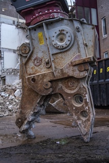 A large, rusted metal demolition claw is positioned on a wet, muddy ground, with crumbling bricks and debris in the background. The claw is part of a heavy machine and exhibits wear from extensive use.