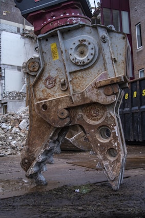 A large, rusted metal demolition claw is positioned on a wet, muddy ground, with crumbling bricks and debris in the background. The claw is part of a heavy machine and exhibits wear from extensive use.