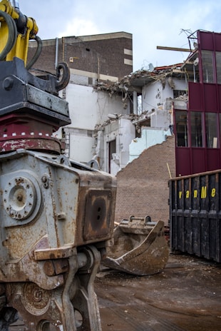 A large piece of heavy machinery is positioned in front of a partially demolished building. The building's exterior shows significant damage, with debris scattered and structural beams exposed. Nearby, a large metal container and a scoop from the machinery are visible.