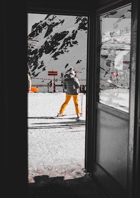 A person dressed in yellow ski pants and a dark jacket stands with skis outside a building, overlooking a snowy mountain landscape. The scene is framed by an open door, creating a sense of looking into a winter sports setting. Snow-covered slopes and rocky outcrops are visible in the background.