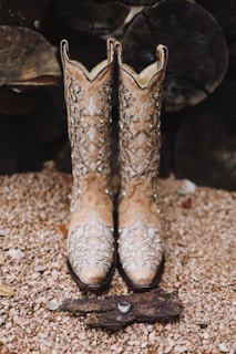 A pair of ornate cowboy boots with intricate designs displayed on a pebbled surface. In front of the boots, there is a piece of wood with a ring placed on it. The background consists of a stack of logs, adding a rustic feel.