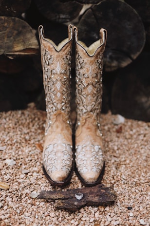 A pair of ornate cowboy boots with intricate designs displayed on a pebbled surface. In front of the boots, there is a piece of wood with a ring placed on it. The background consists of a stack of logs, adding a rustic feel.