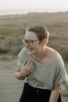 A person with short hair and glasses is laughing in an outdoor setting. The scene is lit by soft, natural light, suggesting sunset or sunrise. The person is wearing a casual gray shirt and appears relaxed and joyful. The background consists of a blurred landscape with earthy tones, possibly a field or coastal area.