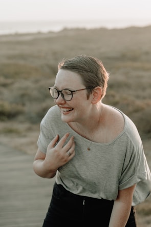 A person with short hair and glasses is laughing in an outdoor setting. The scene is lit by soft, natural light, suggesting sunset or sunrise. The person is wearing a casual gray shirt and appears relaxed and joyful. The background consists of a blurred landscape with earthy tones, possibly a field or coastal area.